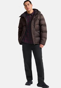 Braune Steppjacke mit Kapuze und gestepptem Design. Trägt darunter ein dunkles kariertes Hemd und schwarze Cargohosen, kombiniert mit dunklen Schuhen.