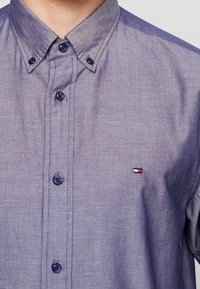 Gładka purpurowa koszula z guzikami i kołnierzykiem zapinanym na guziki, z małym prostokątnym logo w kolorach czerwonym, białym i niebieskim po lewej stronie klatki piersiowej.