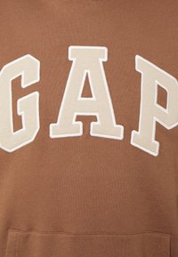 Tessuto marrone con grandi lettere beige "GAP" cucite sopra, che mostrano un'accurata cucitura e texture in una sezione ravvicinata di un indumento.