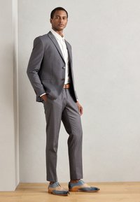 Completo grigio su misura con due bottoni, abbinato a una camicia bianca e una cintura color sabbia. Eleganti scarpe da uomo bicolore con accenti blu e beige.