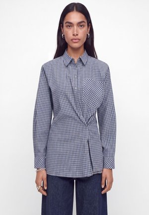Femme aux longs cheveux foncés porte une chemise bleue à carreaux avec un devant asymétrique et une grande poche poitrine, associée à un pantalon bleu foncé.