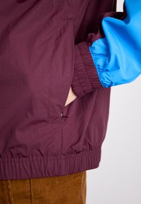 Vindjacka med en vinröd kropp och blå ärmar, med elastiska manschetter och dragkedjefickor på sidorna. Lättviktigt tyg.