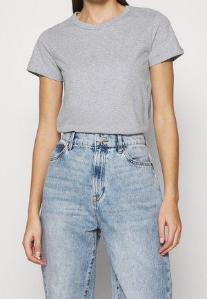 Femme portant un t-shirt gris clair uni à manches courtes, rentré dans un jean en denim bleu clair taille haute, sur un fond uni.