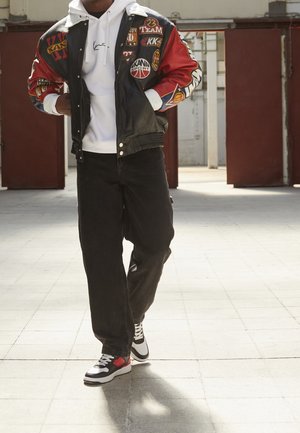 Svart läderbombarjacka med röda ärmar, försedd med patches. Vit hoodie under, kombinerad med lösa svarta jeans och svarta/röda sneakers.