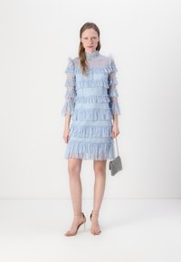 Abito in pizzo azzurro chiaro con volant a strati, maniche lunghe trasparenti e scollo alto. Abbinato a tacchi beige con cinturino alla caviglia e una piccola borsa d'argento.