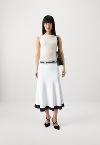 Ermeløs ribbestrikket topp i off-white, parret med en utsvingt hvit skjørt med svart midjebånd og kant. Modellen holder en svart veske og har på seg svarte hæler.