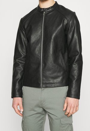 Mann trägt eine eng anliegende schwarze Kunstlederjacke mit Frontreißverschluss und Kragenknopf, kombiniert mit hellgrauen Cargohosen.