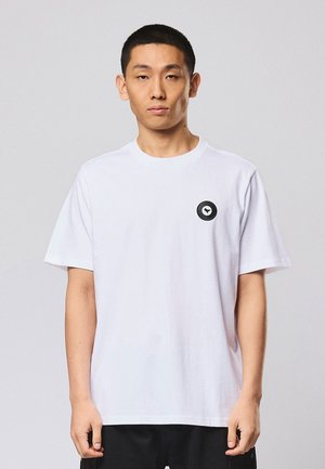 Młody mężczyzna o krótkich włosach nosi prostą białą koszulkę z małym czarnym okrągłym logo na lewej piersi, stoi na tle jasnoszarego tła.