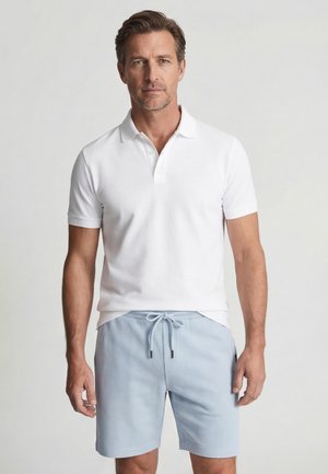 Man draagt een wit poloshirt met korte mouwen en lichtblauwe short met trekkoord, staand tegen een effen lichte achtergrond.