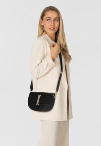 Borsa a tracolla in pelle nera con una texture liscia, hardware in tonalità oro e una forma arrotondata e strutturata. Incluso cinturino regolabile.