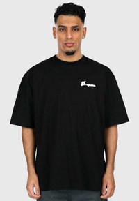 Sort bomulds oversized t-shirt med kort ærme, der har hvidt tekstlogo på venstre bryst. Basis crew neck design, glat tekstur.