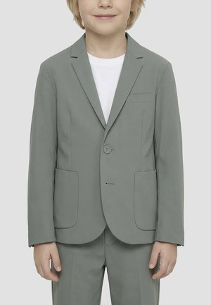 Jeune personne portant un blazer gris clair à deux boutons et un pantalon assorti sur un t-shirt blanc, souriant doucement devant un fond uni.