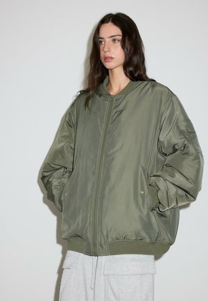 Jeune femme aux longs cheveux foncés portant une veste bomber oversize vert olive et un pantalon gris clair, debout devant un fond clair uni.