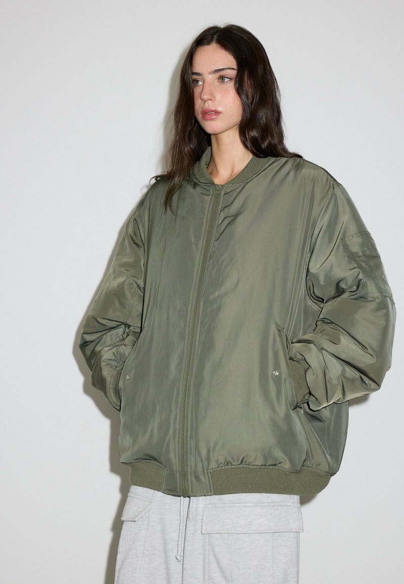 Jeune femme aux longs cheveux foncés portant une veste bomber oversize vert olive et un pantalon gris clair, debout devant un fond clair uni.