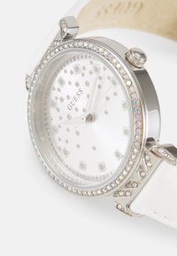 Montre-bracelet argentée avec un bracelet en cuir blanc, un cadran rond, des accents en cristal et un cadran argenté présentant un logo central et des repères horaires carrés.