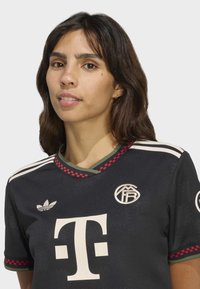 Donna con i capelli scuri che indossa una maglia da calcio nera con strisce bianche, rifiniture a quadri rossi e loghi sul petto e sulle maniche.