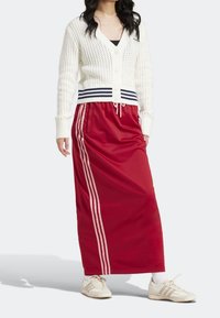 Červená sportovní sukně s bílým šňůrkovým stahováním, s třemi bílými pruhy po stranách. Kombinováno s bílým pleteným svetrem a teniskami.