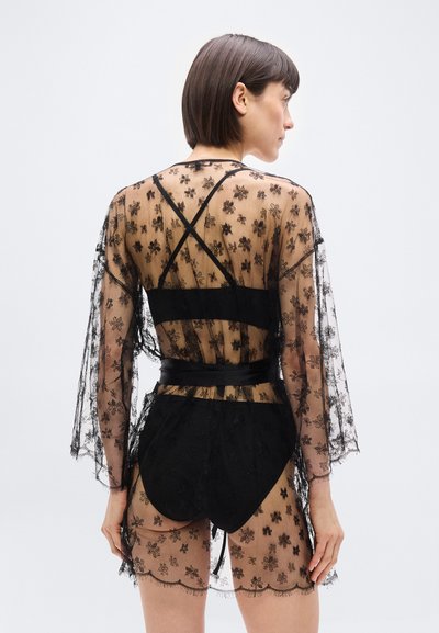 Γυναίκα με κοντά σκούρα μαλλιά, φορώντας διάφανο μαύρο φόρεμα από δαντέλα με floral σχέδιο και μακριά μανίκια, με μαύρα εσώρουχα, γυρισμένη πλάτη σε μια απλή φόντο.