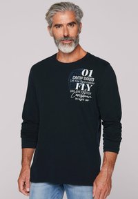 Zwarte long sleeve T-shirt met witte en blauwe grafische tekst op de bovenkant van de voorkant, gemaakt van zachte stof met een relaxte pasvorm. Blauwe stikselaccenten.