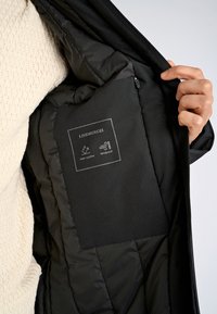 Svart puffjacka med insida som har en vattenavvisande och vindskyddande etikett. Mjuk foder och quiltad struktur synlig runt kanterna.