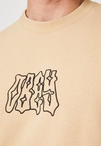 Béžové bavlněné tričko s černým, zkresleným textovým designem na přední straně. Kolo výstřih s viditelnými švy.