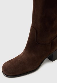 Stivale alla caviglia in suede marrone con punta quadrata, dettagli di cucitura discreti e tacco a blocco. Design elegante con texture liscia.