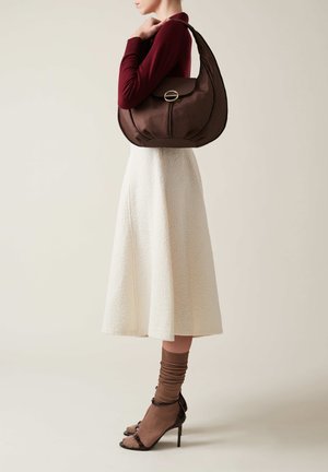 Braune Leder-Schultertasche mit einem runden Design, strukturiertem Oberflächenmaterial und goldfarbenen Beschlägen. Das Model trägt einen weißen Rock und braune Socken mit Absatzschuhen.