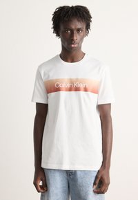 Bílá bavlněná tričko s krátkým rukávem, které má gradientní oranžový pruh a nápis "Calvin Klein" v tučných bílých písmenách přes hruď.