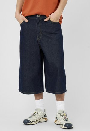 Personne portant un long short en denim foncé, des chaussettes blanches, des chaussures de sport beige et noir, et une chemise orange avec les mains dans les poches.