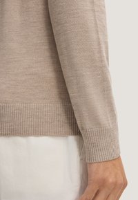 Dettaglio di un maglione lavorato a maglia marrone chiaro, con un polsino e un orlo a coste e una texture liscia. Sotto, un indumento bianco è parzialmente visibile.