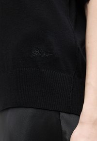 Μάλλινο μαύρο πουλόβερ με διακριτικό κεντητό λογότυπο στη ραφή, σε συνδυασμό με μαύρο, λείο παντελόνι, και εμφανές αντιβράχιο στη δεξιά πλευρά.