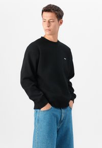 Črna majica s kapuco z okroglim izrezom, ki ima rebraste manšete in majhen bel logo na prsih. Uporabljena z svetloblue širokimi kavbojkami.