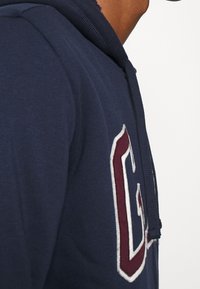 Tmavě modrá mikina s kapucí, vybavená přední kapsou, stahovací šňůrkou na kapuci a výšivkou písmen ve vínové a bílé barvě na hrudi. Jemná textura látky.