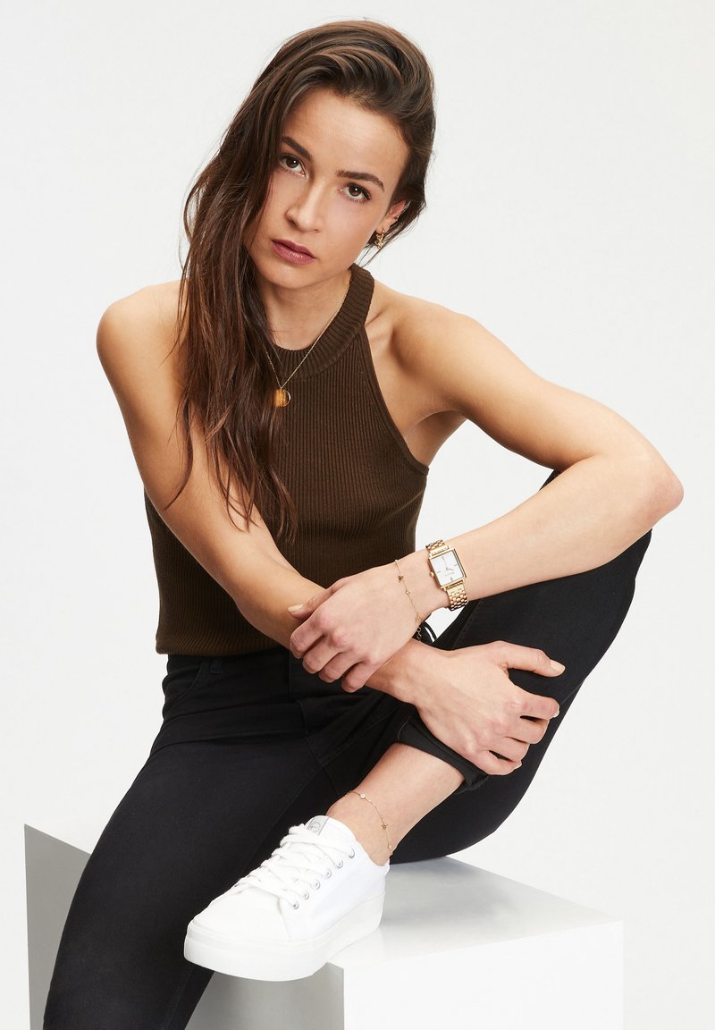 Braun gestreiftes Tanktop, schwarze eng anliegende Jeans, weiße Plateausneaker. Goldene Uhr und Armbänder an den Handgelenken, sitzende Pose mit einem angewinkelten Bein.