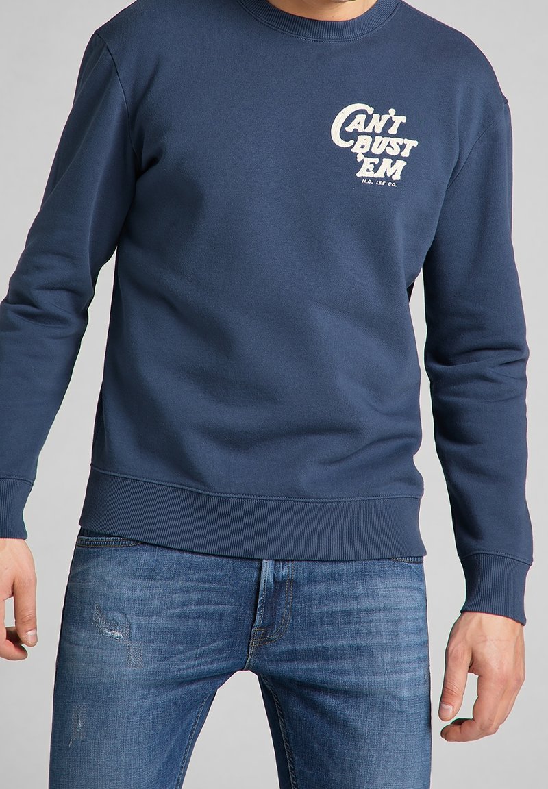 Marinblå sweatshirt med rund halsringning, ribbade muddar och nederkant, med vit textgrafik som säger "KAN INTE SLÅ DEM" på vänster bröst. Denimmer.