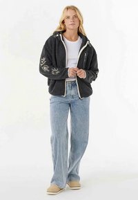 Schwarzer Fleecepullover mit weißer Blumenstickerei, Reißverschluss und verstellbaren Kordeln. Kombiniert mit hellblauen Schlagjeans und beigen Schuhen.