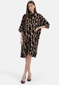 Schwarz-beiges gemustertes Oversized-Hemdkleid mit kurzen Ärmeln, Druckknopfverschluss und lockerer Passform, kombiniert mit schwarzen Absätzen.