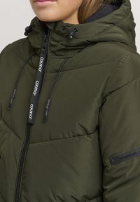 Padded olive green jacket with a high collar, adjustable hood, black zipper, and side pocket featuring a zip closure.