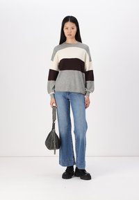 Pull rayé gris, crème et bordeaux ; coupe ample, texture côtelée ; associé avec un jeans évasé et des chaussures noires chunky ; tenant un sac à main sombre.