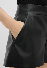 Pantaloni shorts neri in finta pelle con vita alta e tasche laterali. La texture è liscia con una leggera lucentezza. Design a fronte piatto.