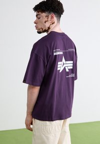 Lilla bomulds t-shirt med korte ærmer, med et hvidt grafisk design på ryggen og et lille mærke på ærmet. Enkelt på fronten.