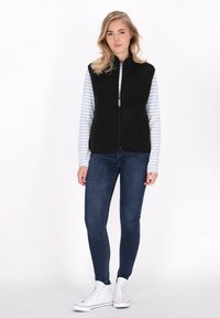 Melns flīsa veste ar rāvējslēdzēju, uzvilkta virs zilā un baltā svītrainā garām piedurknēm t-kreklā, saskaņota ar tumšām džinsām un baltām sporta apaviem.