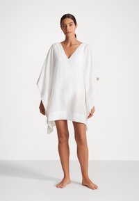 Balts lina kaftans ar V veida kakla izgriezumu, platiem piedurknēm un asimetrisku apakšmalu. Mīksta tekstūra, viegls audums un minimālisma dizains.