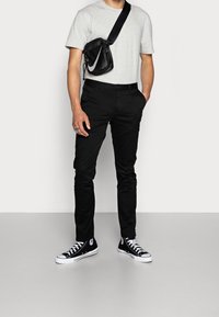 Pantaloni neri slim-fit, t-shirt grigia a collo tondo e sneakers nere con dettagli bianchi. Borsa a tracolla nera con un dettaglio di design bianco.