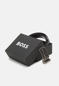 Čierny kožený opasok s obdĺžnikovou striebornou kovovou prackou, prezentovaný v čiernej škatuľke s nápisom "BOSS" vytlačeným bielou farbou.