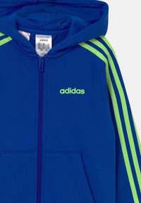 Sudadera azul con capucha que presenta una cremallera completa, acentos verdes y tres rayas verdes a lo largo de las mangas. Logotipo "adidas" en verde en el pecho.