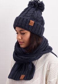 Navy knit beanie with pom-pom and leather tag, paired with a matching textured scarf. Both have ribbed patterns and thick yarn.
