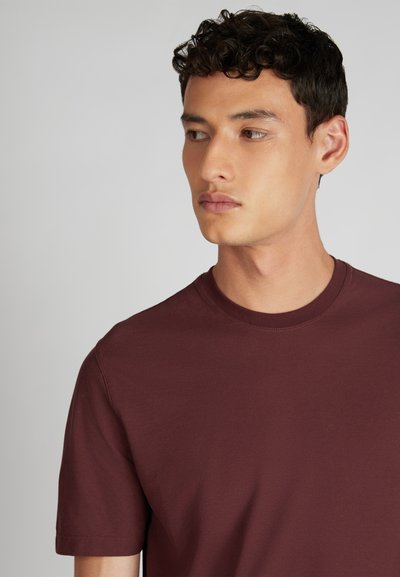 Jeune homme à cheveux courts et bouclés, de couleur noire, portant un t-shirt uni bordeaux, regardant à sa gauche sur un fond gris clair.