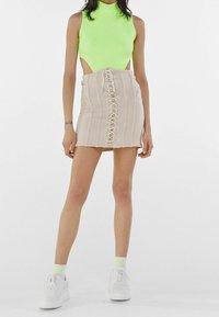 Top sans manches vert néon associé à une mini jupe beige à rayures verticales et un détail de lacet au centre ; complété par des baskets blanches.