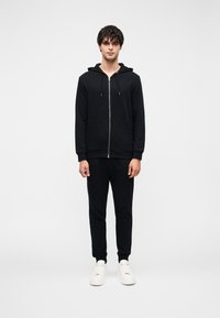 Μαύρο φούτερ με φερμουάρ, κουκούλα με κορδόνι και μπροστινές τσέπες, σε συνδυασμό με μαύρη φόρμα που ταιριάζει και λευκά αθλητικά παπούτσια. Ομαλή υφή.